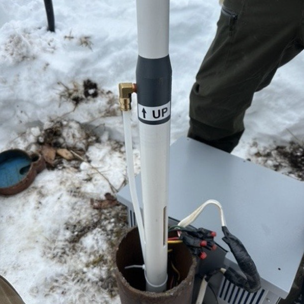 A close-up of an Airwell radon mitigation system installation in a snowy outdoor environment. The image shows a white PVC pipe labeled "UP" connected to a device with electrical wiring and tubing visible. The system is designed to extract radon gas from the ground, helping improve indoor air quality.